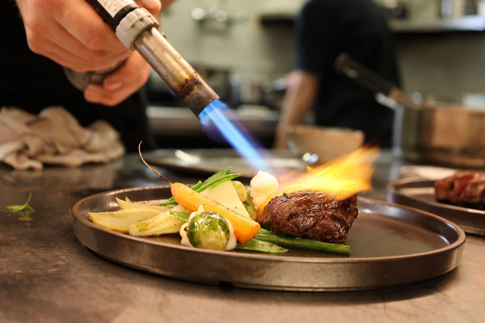 Chef blowtorching a plated steak with vegetables at the Two Hat Venue placement